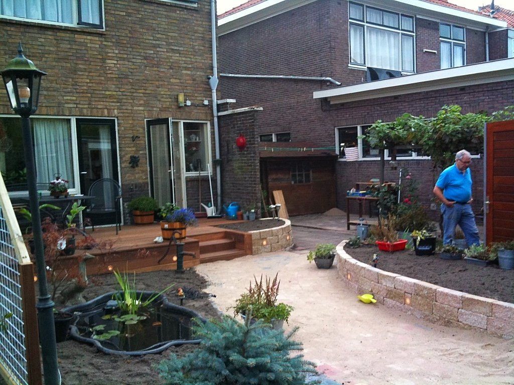 Achtertuin met een man bij een bloembed, pad, vijver, bakstenen huis, bruine bakstenen muren en een lantaarn.