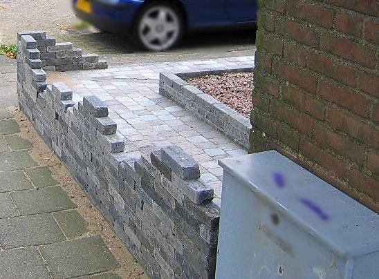 Een stenen keermuur naast een stenen pad en een gebouw. ​​Op de achtergrond staat een blauwe auto geparkeerd.