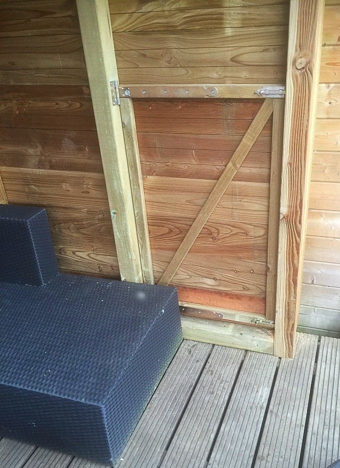 Een houten poort, open, binnen een met houten panelen beklede constructie. Er staat een blauwe buitenbank voor.