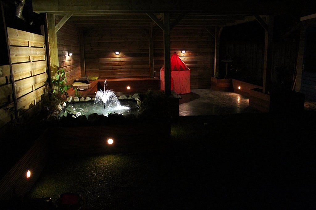 Een nachtelijke buitenscène met een verlichte waterfontein, een houten constructie en kleine grondlampjes.