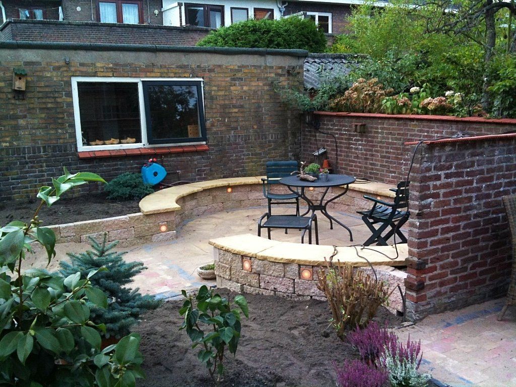 Gebogen terras met tafel en stoelen, omgeven door bakstenen muren. Een tuin met planten omringt het terras.