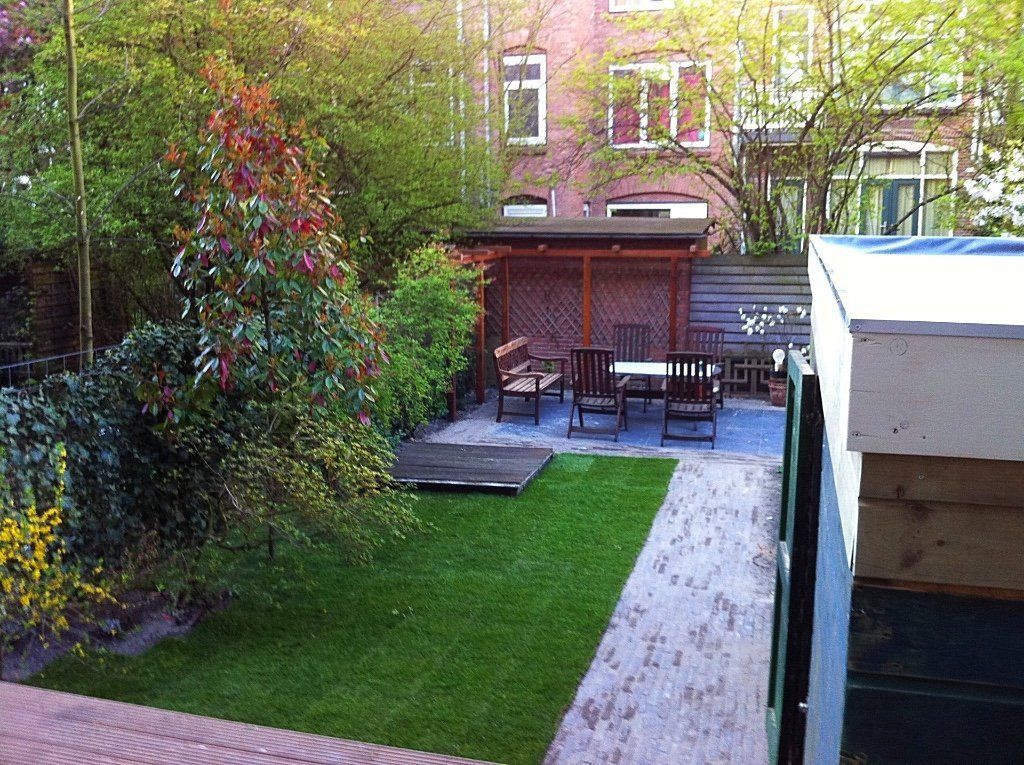 Achtertuin met terras, tafel, stoelen, pergola en groen gazon, met gebouwen op de achtergrond.