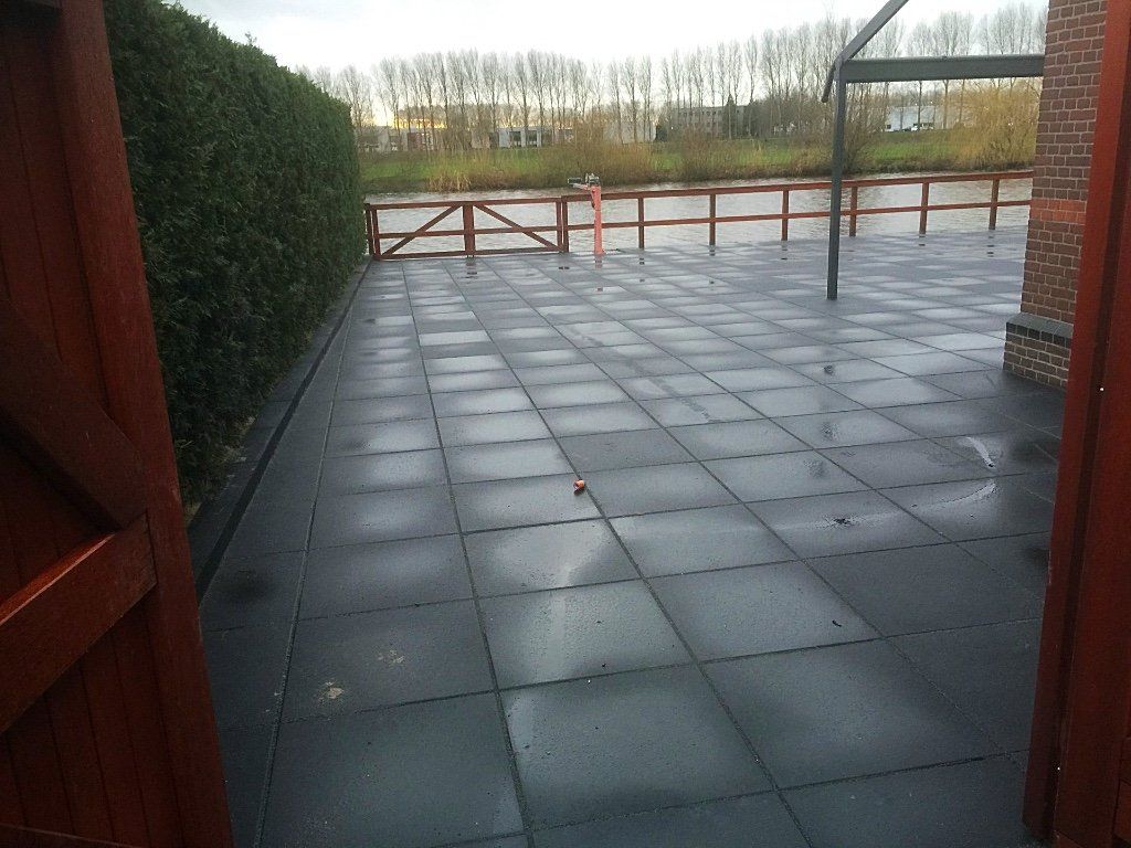 Terras met donkere vierkante tegels, een haag, een houten schutting en water op de achtergrond.