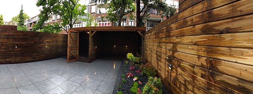 Een houten schuur, een pad en muren vormen een contrast met een woonomgeving met bomen.