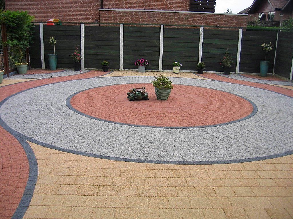Rond terras van bakstenen met concentrische ringen van rode, grijze en gele bakstenen; potplanten, groene schutting op de achtergrond.