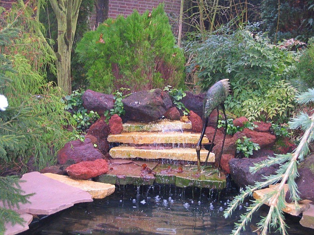 Een kleine, trapsgewijs oplopende waterval die in een vijver uitmondt, omgeven door rotsen en groen.