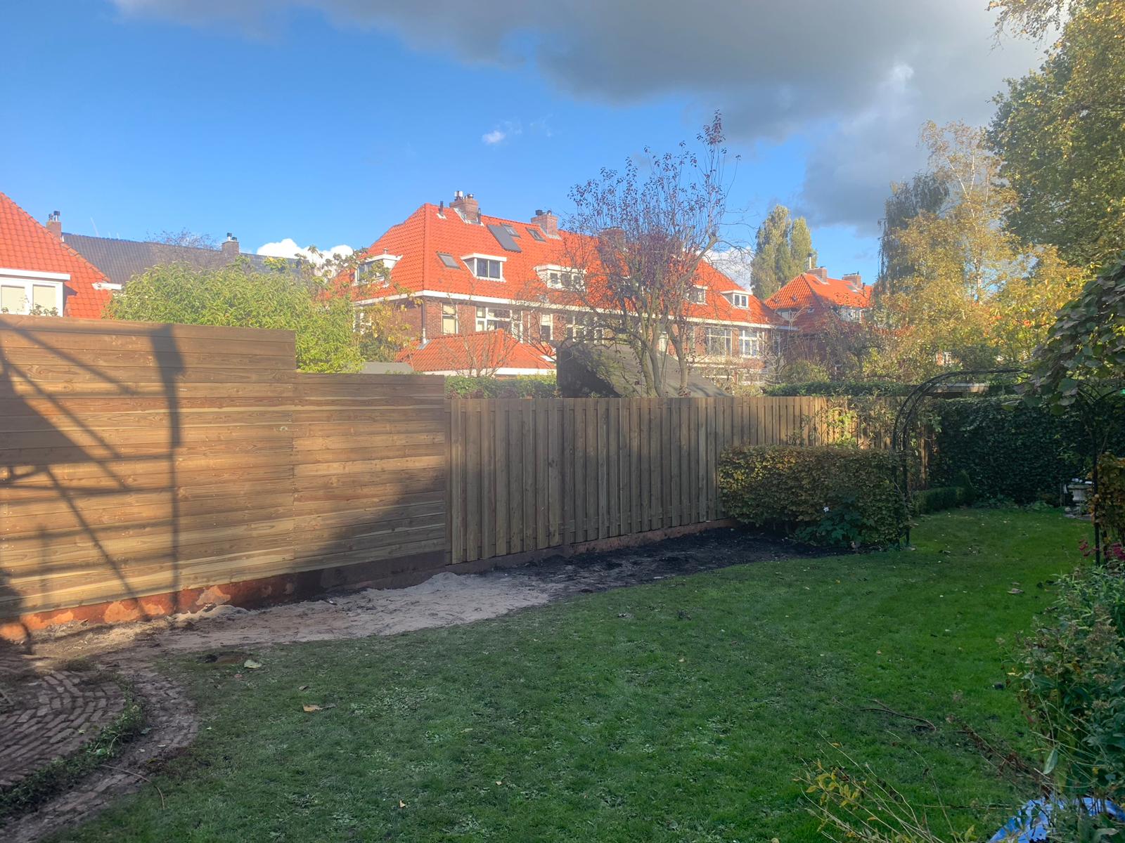 Houten schutting in de achtertuin met gras, een smal grindpad en huizen op de achtergrond.
