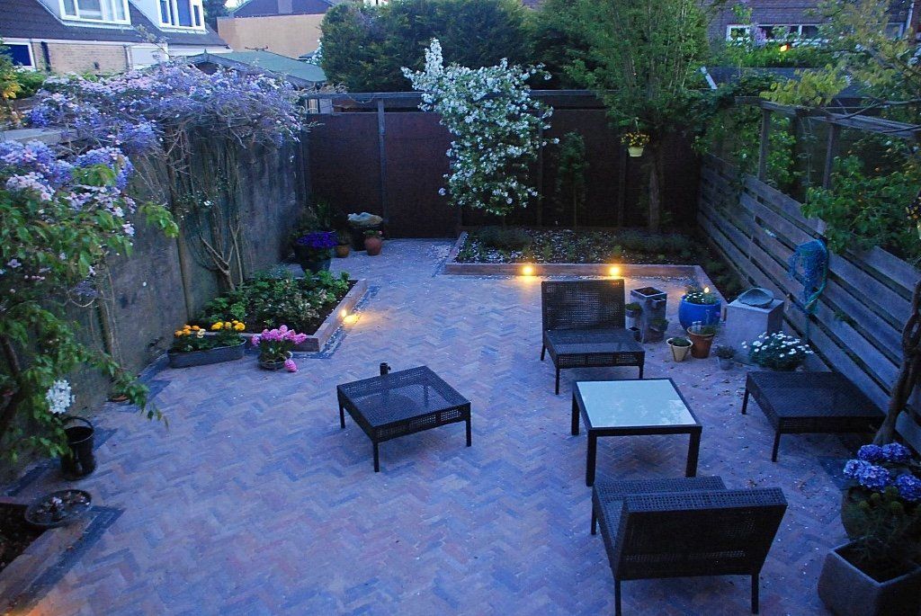 Een betegelde achtertuin met terrasmeubilair, planten en sfeerverlichting in de schemering.