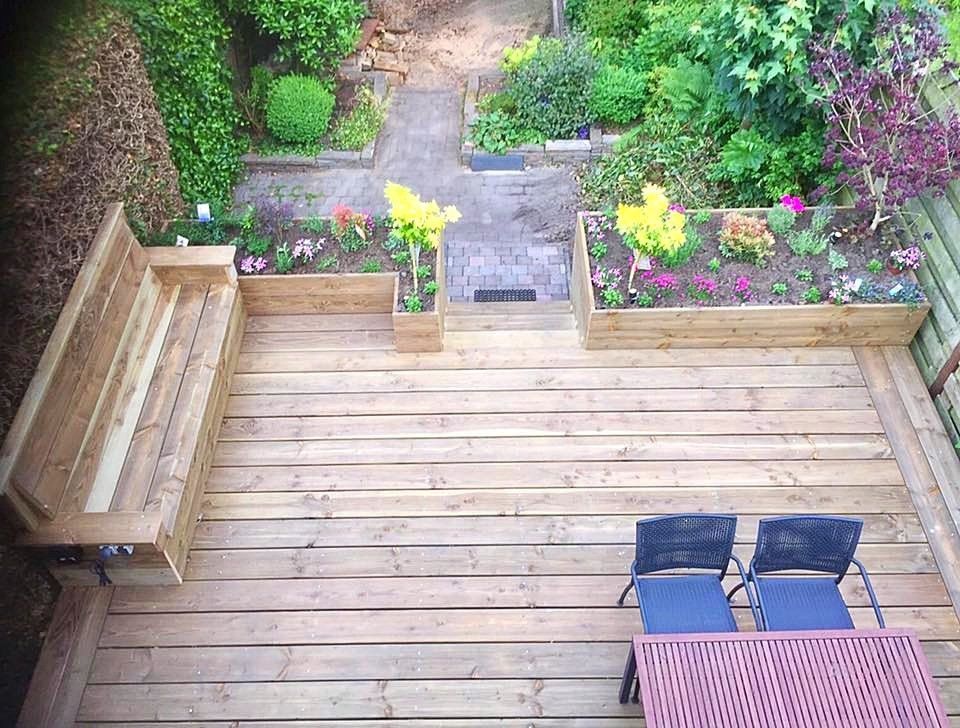Bovenaanzicht van een houten terras met ingebouwde zitgelegenheid, bloembakken en een smal tuinpad. Op het terras staan ​​twee stoelen en een klaptafel.