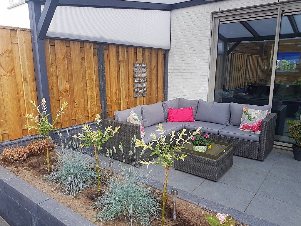 Terras buiten met een grijze hoekbank, salontafel en planten tegen een houten schutting.