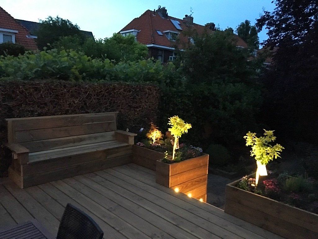 Houten terras met bankje en verlichte plantenbakken in de schemering. Huizen en begroeiing op de achtergrond.