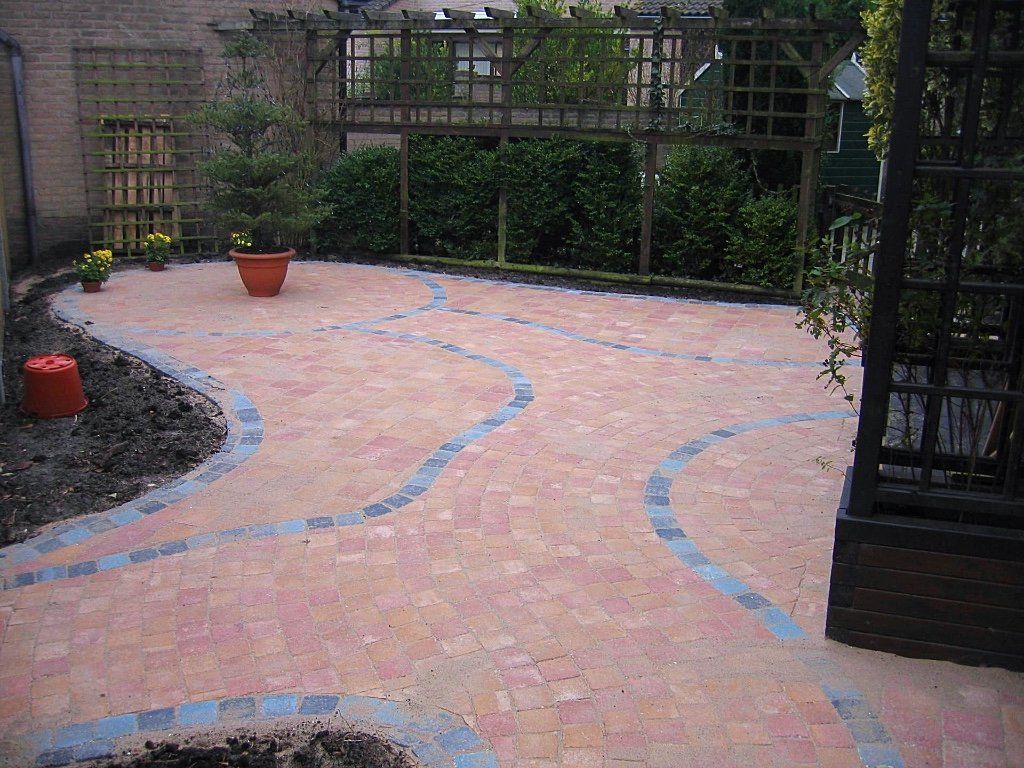 Een bakstenen terras met kronkelende paden van blauwe natuursteen, omgeven door planten en een houten pergola.