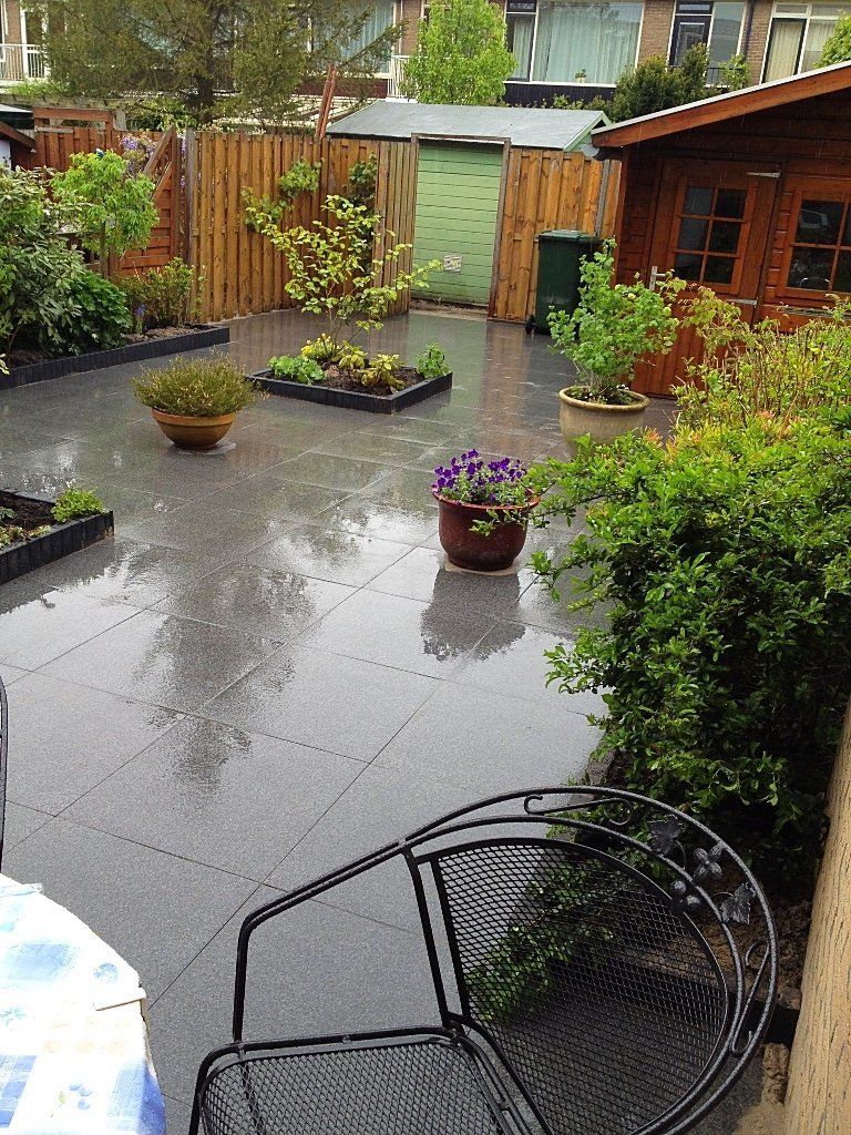 Terras in de achtertuin met natte grijze tegels, planten in potten, houten schuur, schutting en metalen stoel.