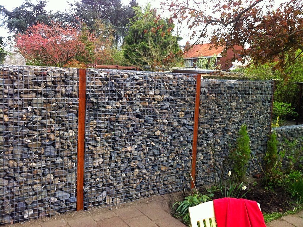 Gabionstenen schutting met bruine metalen steunen in een tuinomgeving.