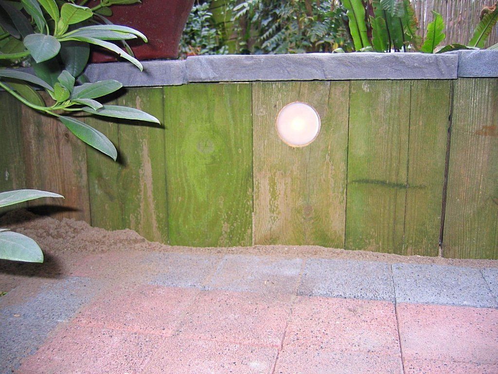 Een verweerde houten muur met een ronde lamp, omgeven door planten en bakstenen bestrating.