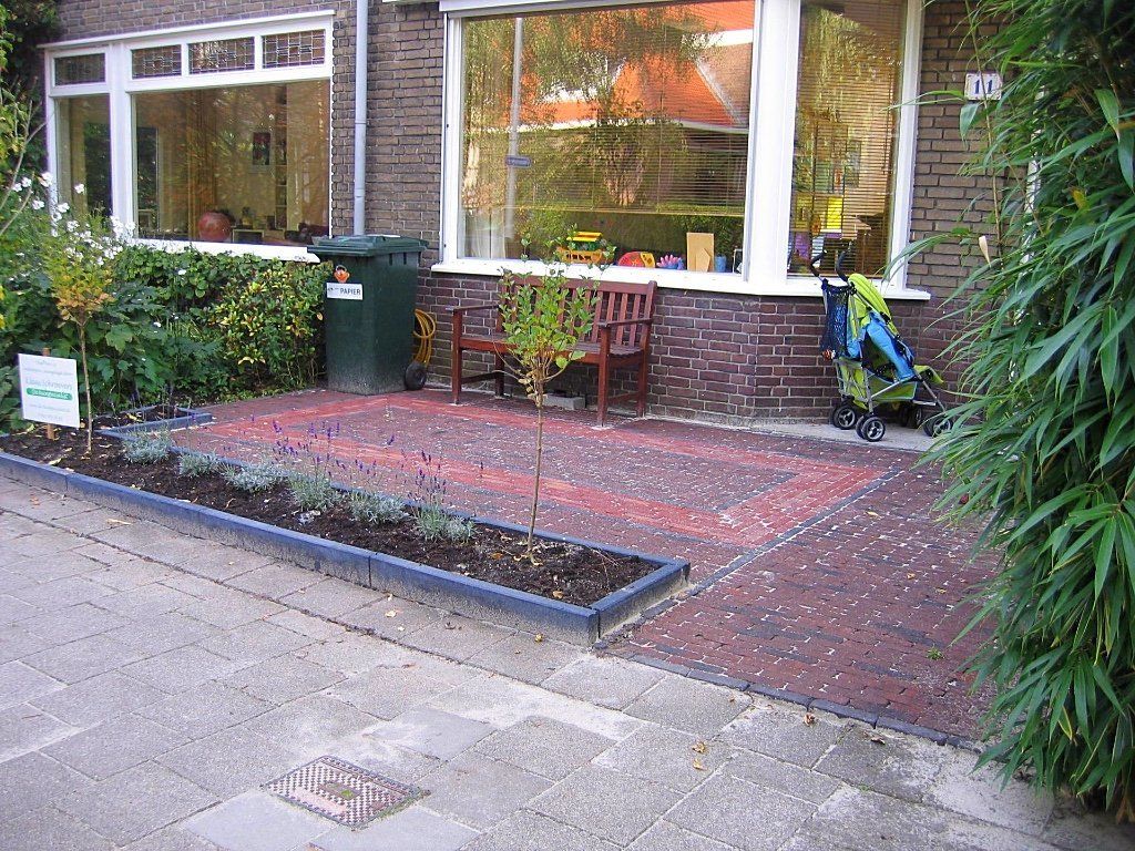 Een stenen terras met een kleine tuin voor een huis. Een bankje en een kinderwagen staan ​​bij de ramen.
