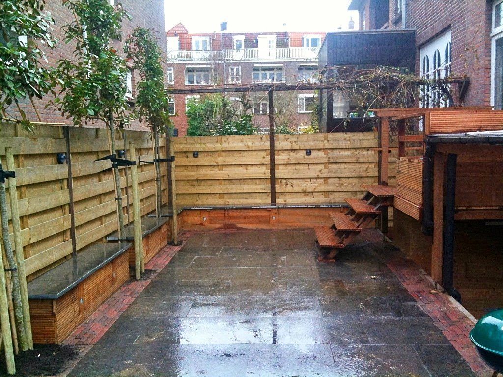 Houten schutting in de achtertuin met een terras van bakstenen, trappen en kleine bomen. Bewolkte lucht.