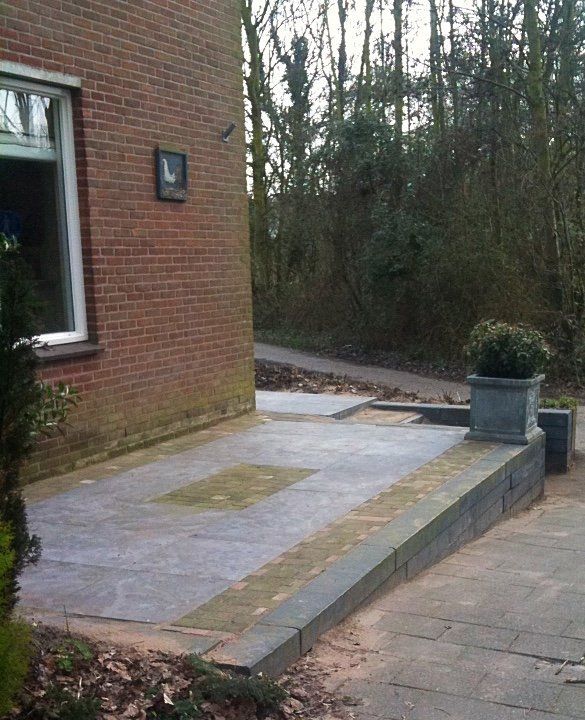 Bakstenen gebouw met een betonnen terras. Bloembak tegen een laag muurtje. Pad leidt naar bomen.