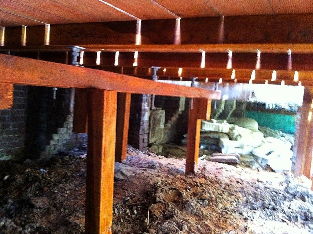 Binnenruimte onder het huis met houten balken en bakstenen fundering. Aarden vloer, veel licht.