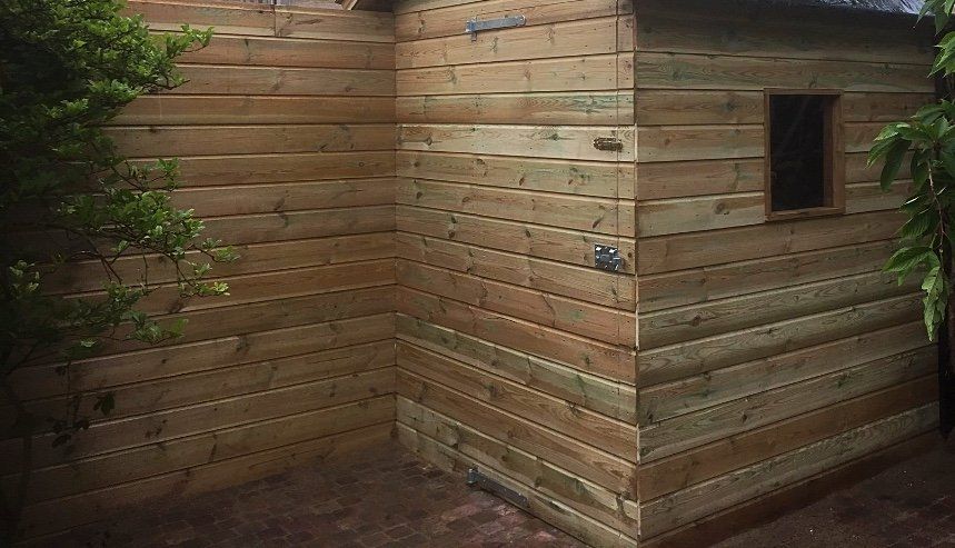 Houten schuur met horizontale planken als gevelbekleding, een klein raam en omgeven door begroeiing.