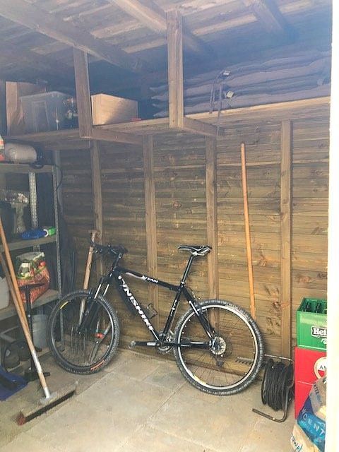 Een zwarte fiets staat in een houten schuur tegen de muur. Op de planken erboven staan diverse spullen.