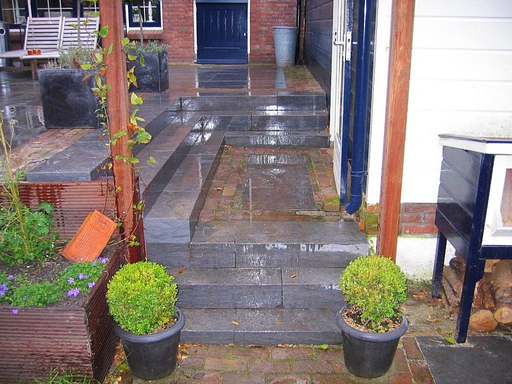 Stenen trappen leiden naar een donkerblauwe deur, geflankeerd door potplanten, en een houten pergola aan de linkerkant.