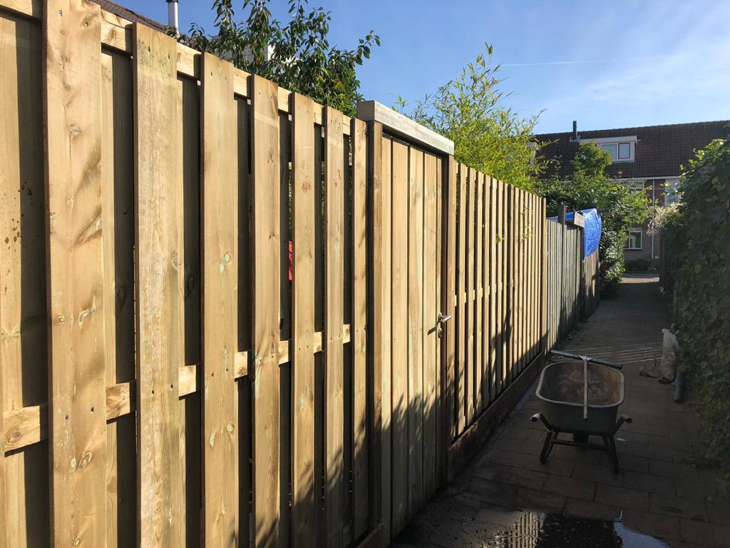 Houten schutting in een achtertuin met een poort en een kruiwagen op een verhard pad.