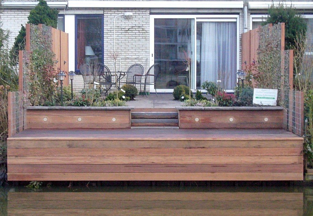 Houten terras met ingebouwde plantenbakken, met uitzicht op het water, voor een gebouw met glazen deuren.