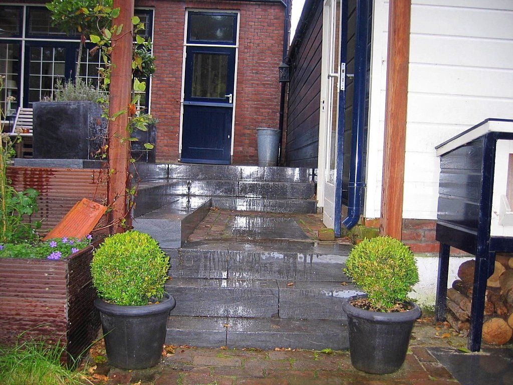 Stenen trappen leiden naar een donkerblauwe deur tussen gebouwen, geflankeerd door groene struiken in potten.