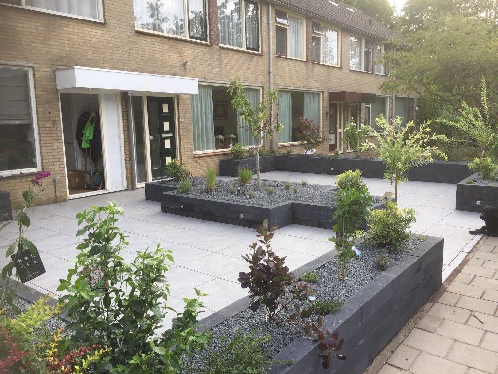 Grijze patio met verhoogde plantenbakken gevuld met donkere stenen en jonge bomen voor een gebouw.