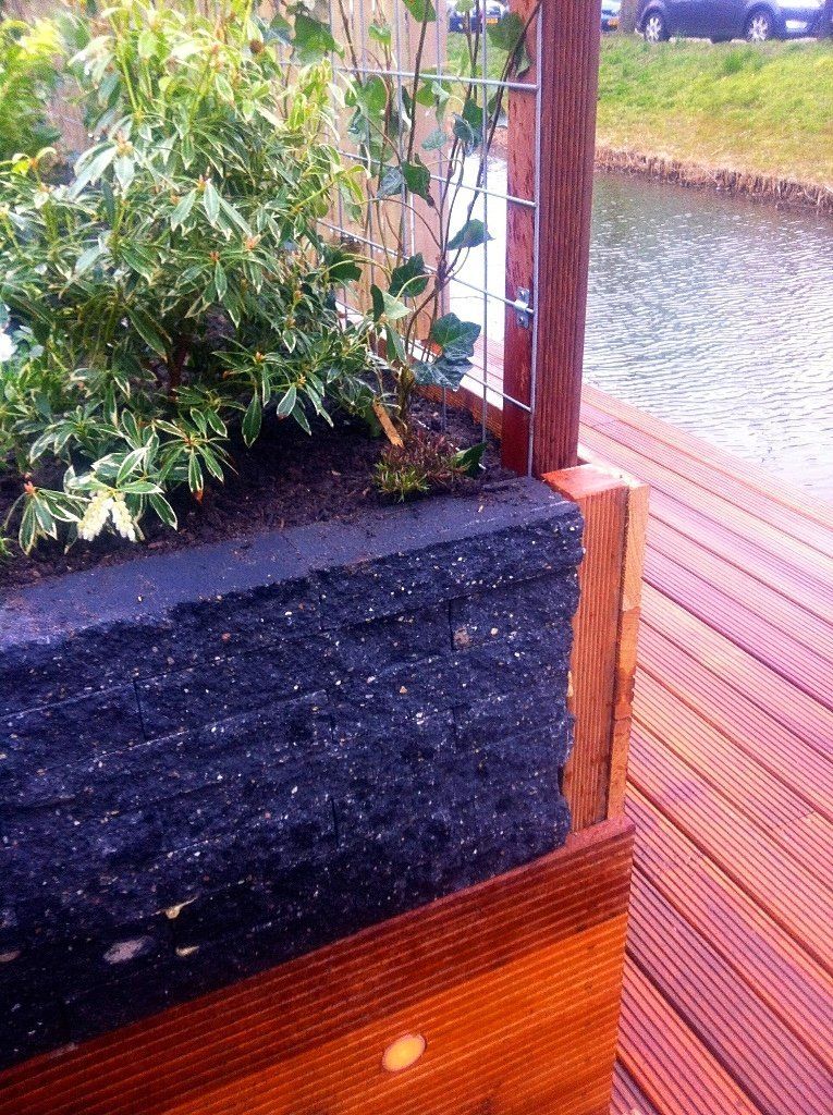 Bloembak van zwart gerecycled materiaal, hout en een metalen klimrek, vlakbij water.