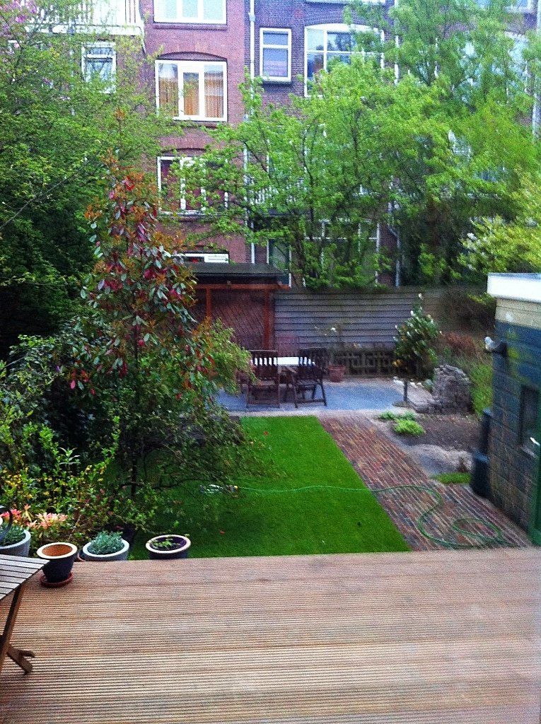 Achtertuin met gazon, terras, tafel, schuur en bakstenen gebouwen op de achtergrond.