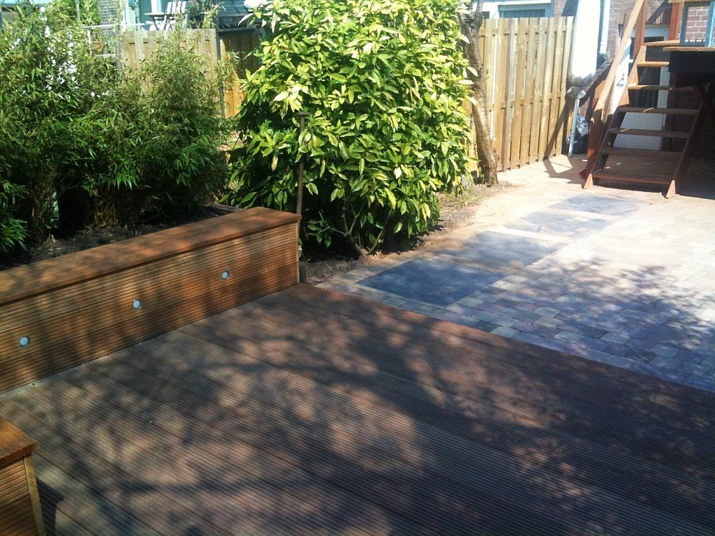 Terras met houten vlonder, ingebouwde bank, bestraat pad en weelderige groene struiken, zonnige dag.