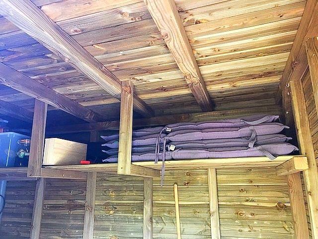 Een houten opbergrek in een schuur, met opgerolde paarse matten erop. Het dak en de muren van de schuur zijn ook van hout.