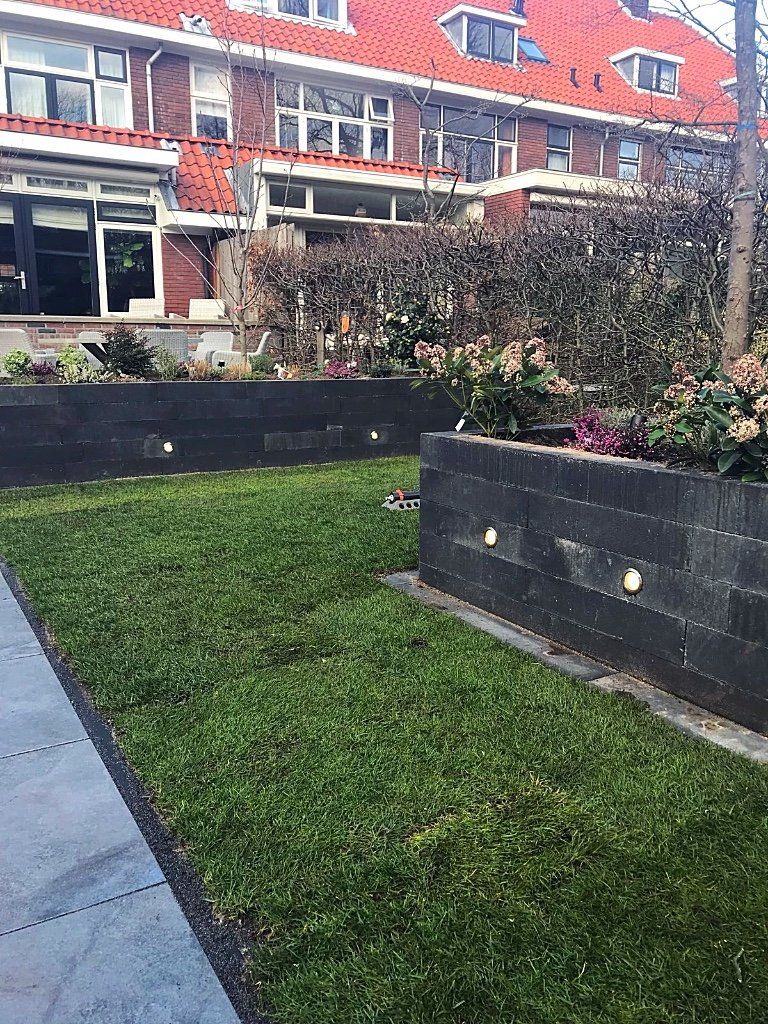Een groen gazon tussen donkergrijze blokmuren met ingebouwde verlichting, planten en op de achtergrond gebouwen met rode daken.