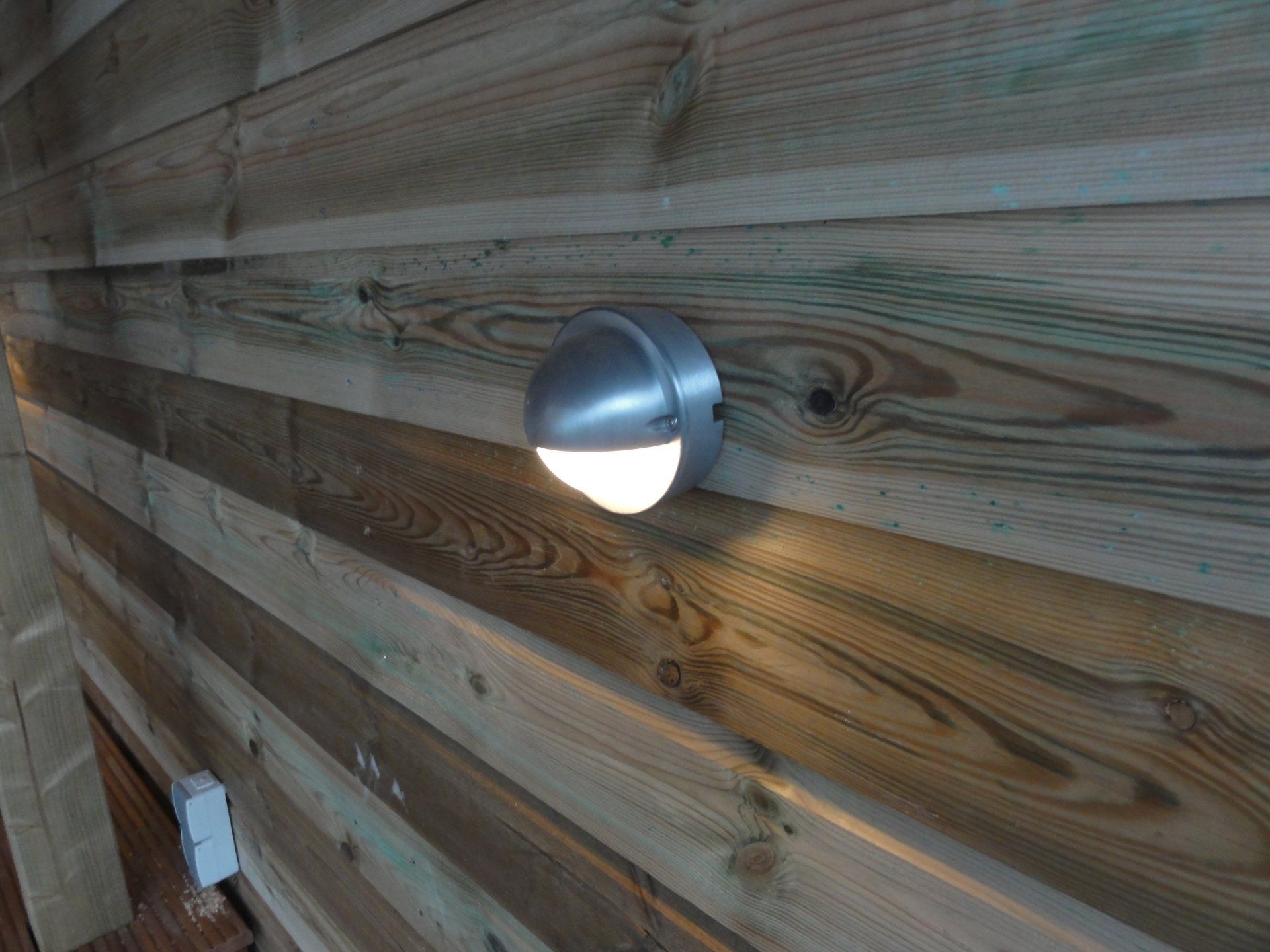 Een zilverkleurige buitenwandlamp is gemonteerd op horizontale houten planken en verlicht het oppervlak.