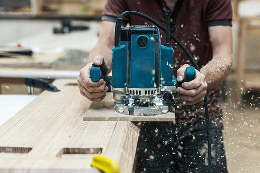Een persoon gebruikt een blauwe freesmachine op een houten plank om houtkrullen te maken in een werkplaats.