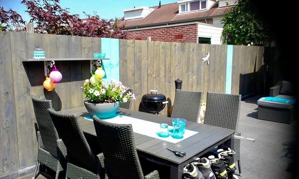 Buitenpatio met grijze tafel, stoelen en barbecue tegen een houten schutting met turquoise accenten.