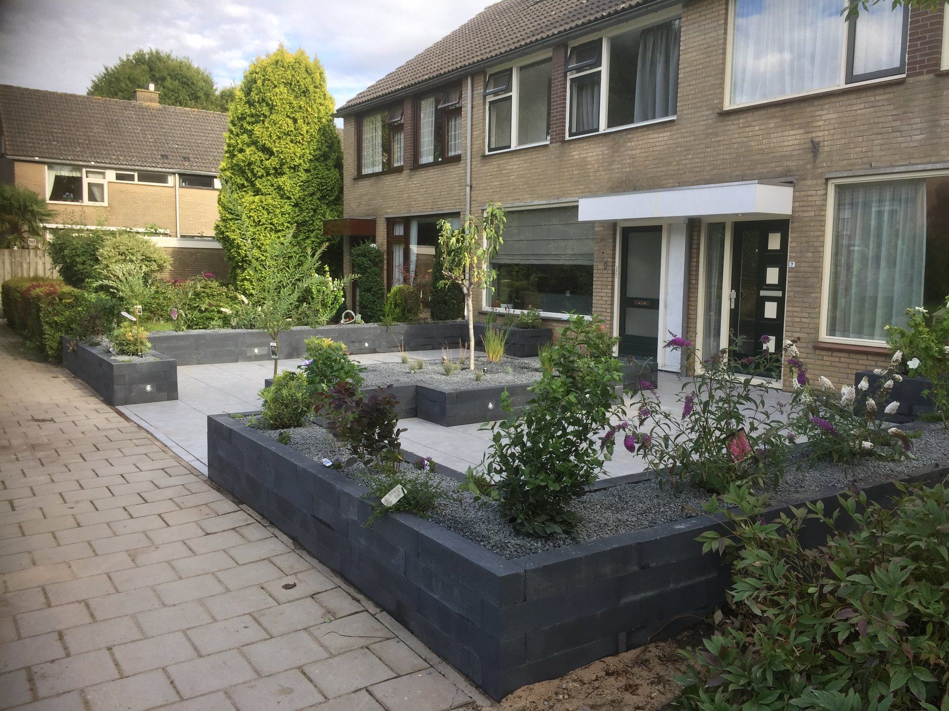 Een huis met een bakstenen gevel en een moderne, aangelegde voortuin met verhoogde plantenbakken en een betegeld terras.