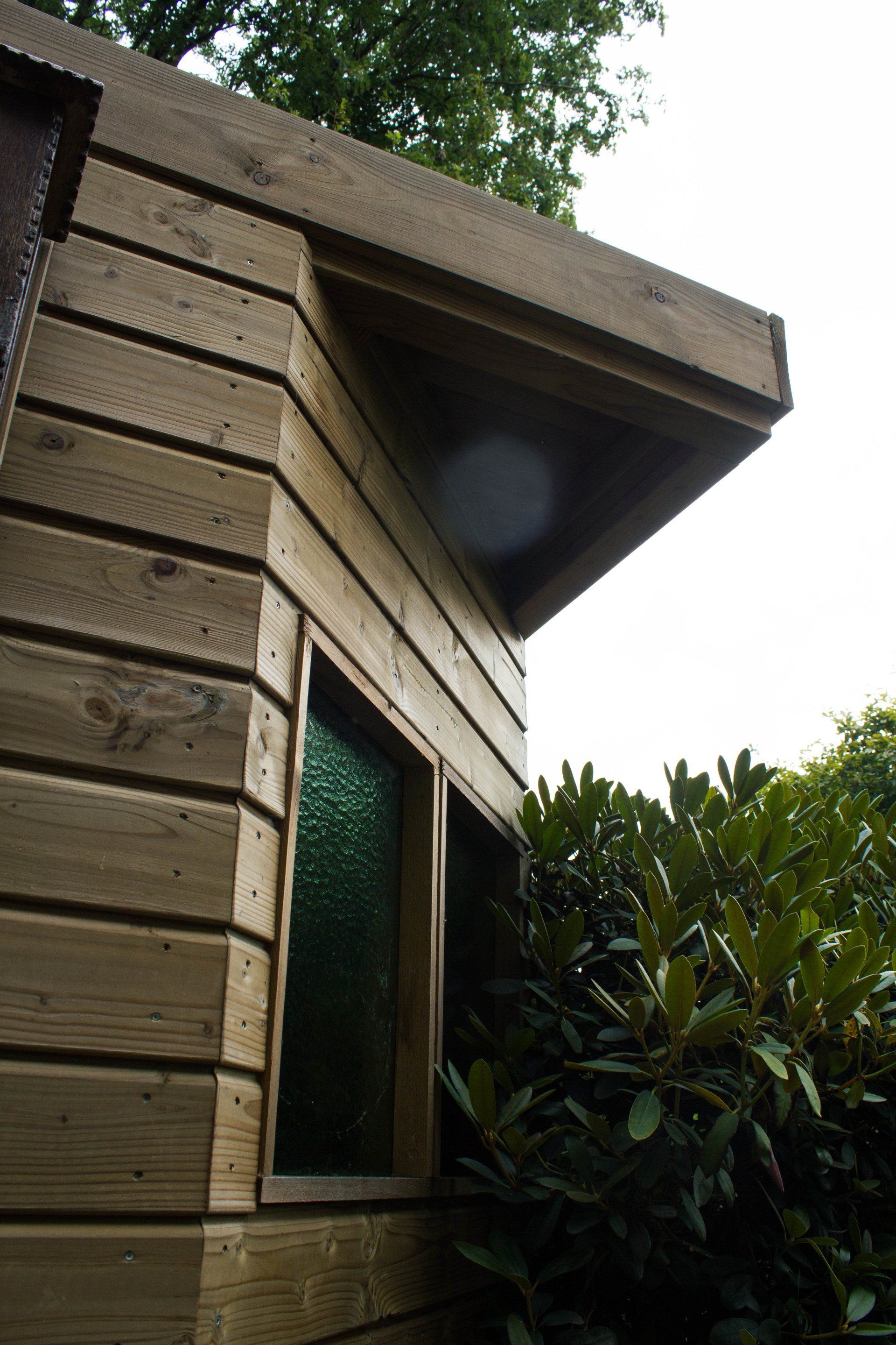 Houten hoekgebouw met raam, groen glas en overhangend dak. Donkere houten gevelbekleding en begroeiing.