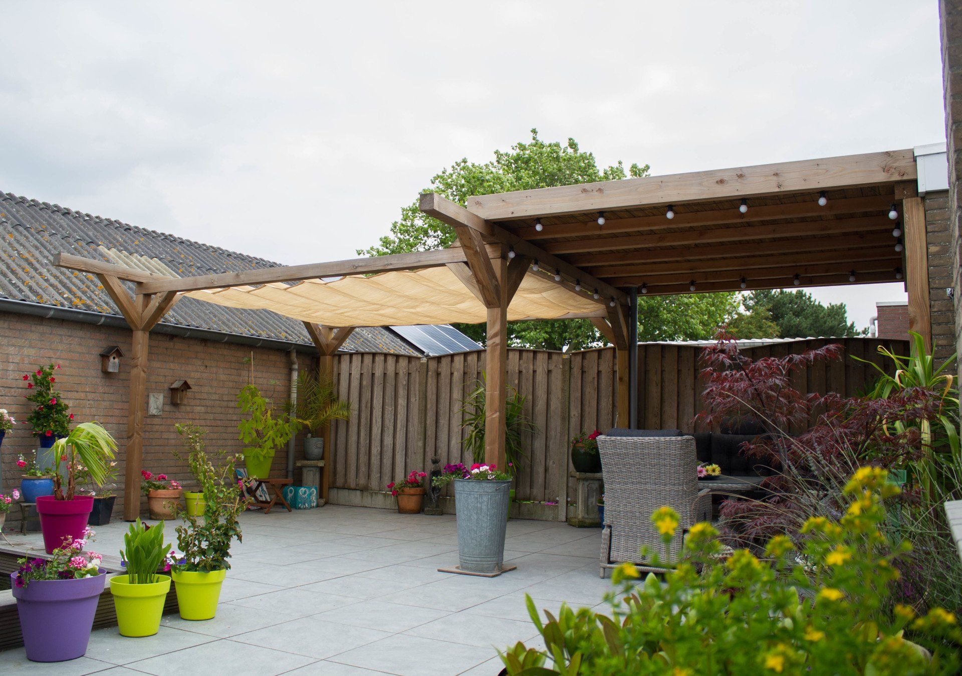 Terras met houten pergola, grijze stenen bestrating, potplanten en houten schutting.