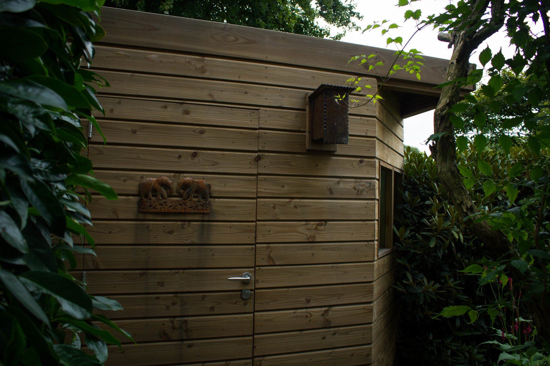 Houten schuur met een vogelhuisje en een plaquette, omgeven door groen.