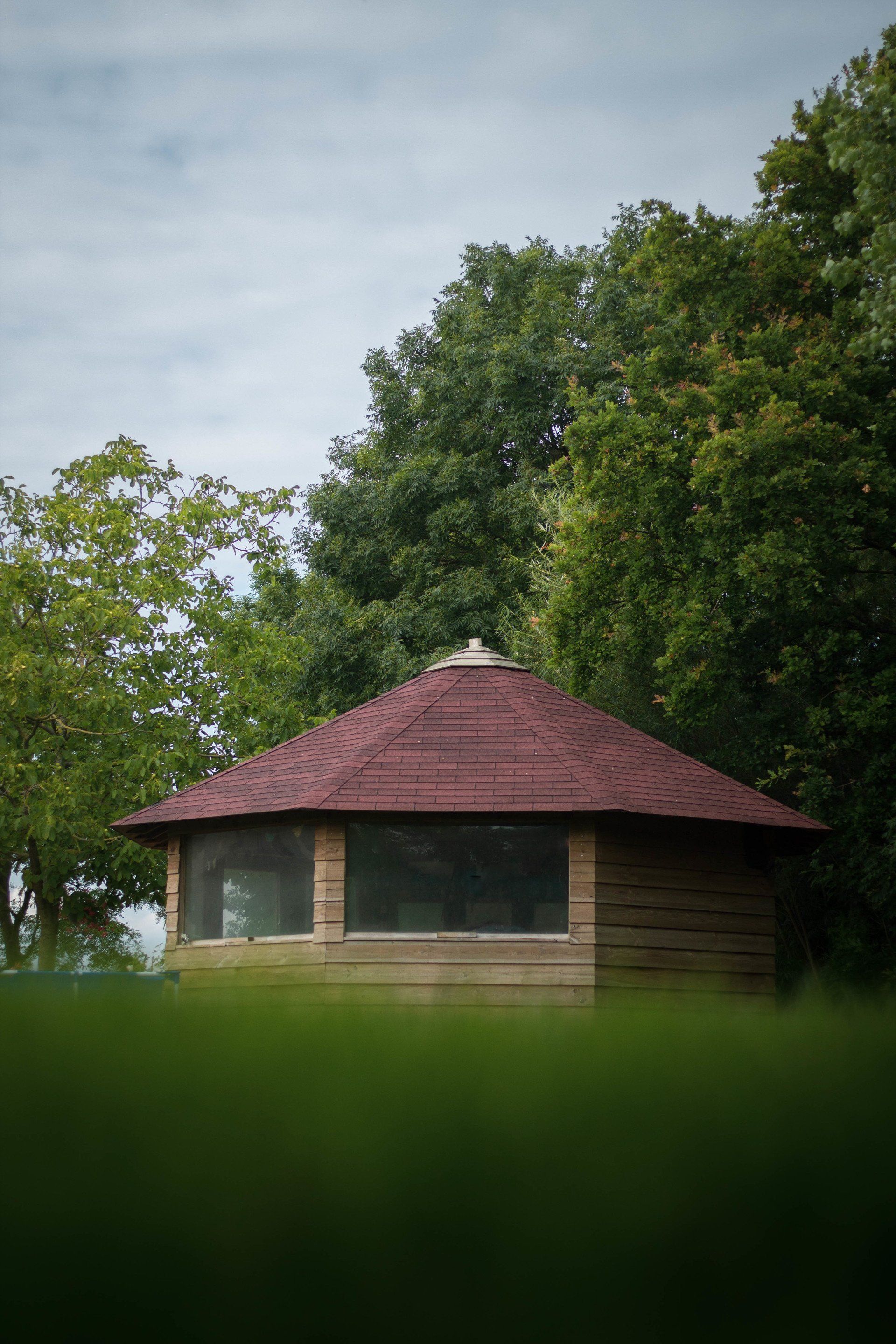 Houten prieel met rood dak in een groen grasveld, omgeven door bomen, onder een bewolkte hemel.