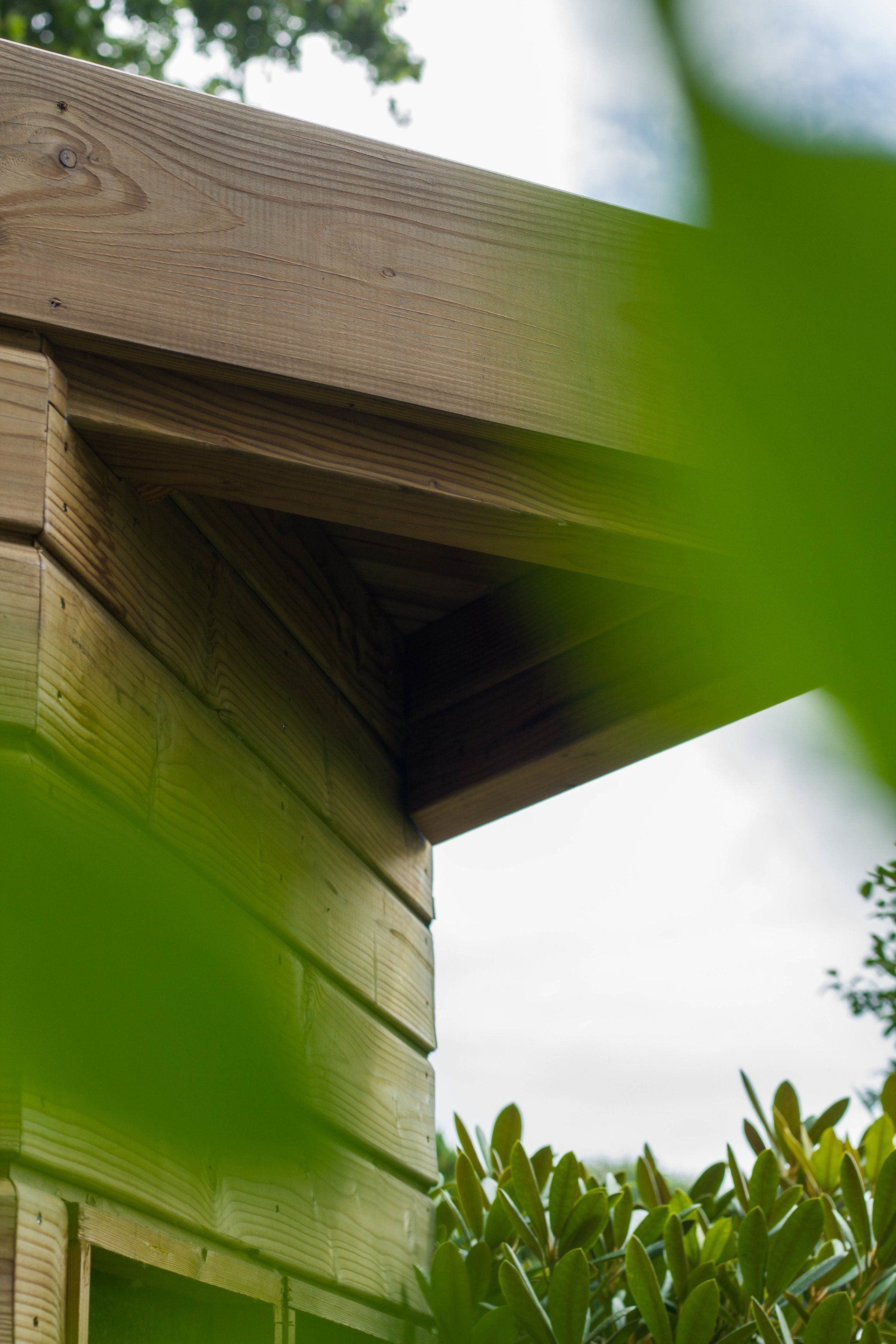 Hoek van een houten gebouw met een overhangend dak, zichtbaar door het groene gebladerte.
