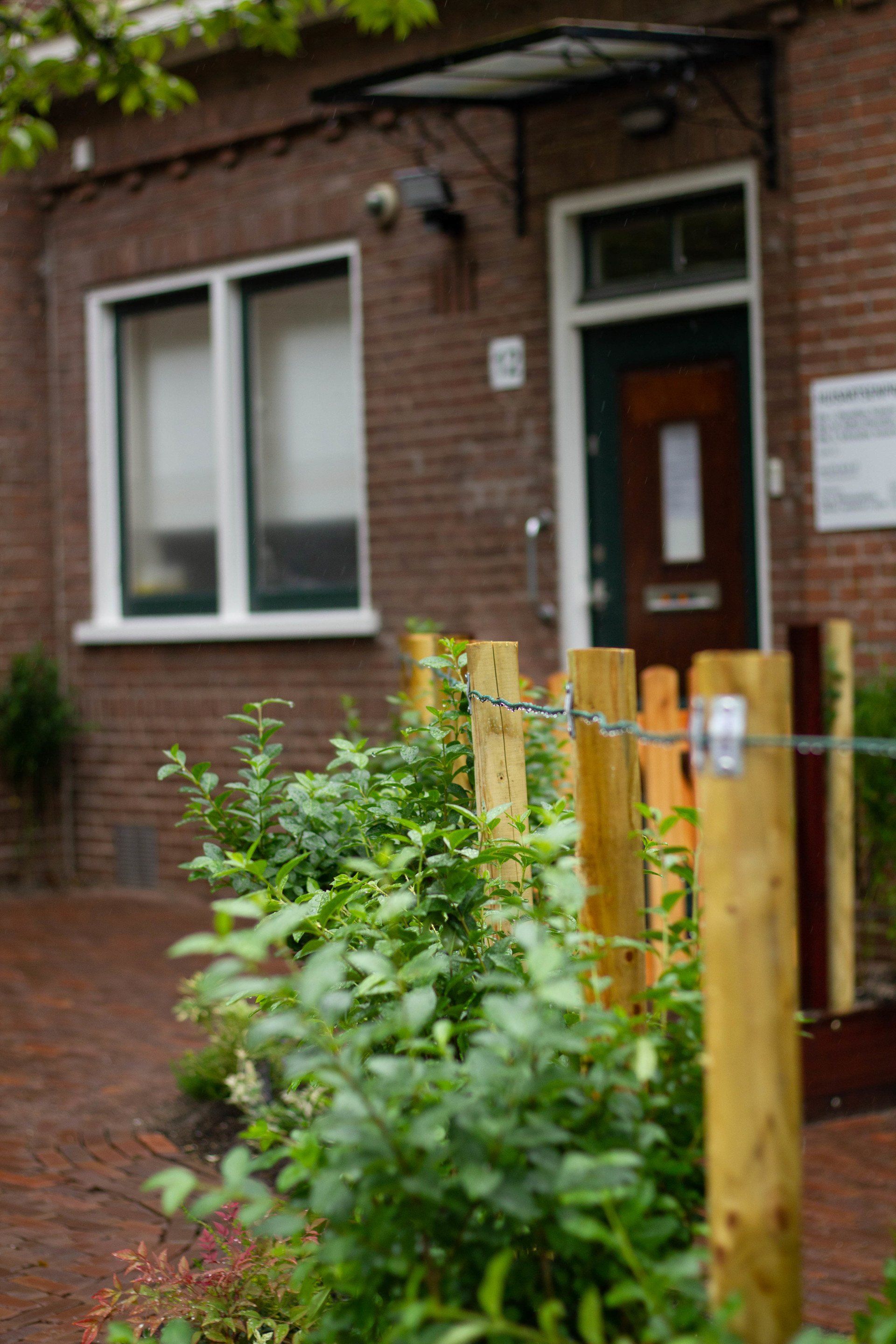 Bakstenen gebouw met houten palen, draadhek en groen ervoor. Deur met zichtbaar glaspaneel.