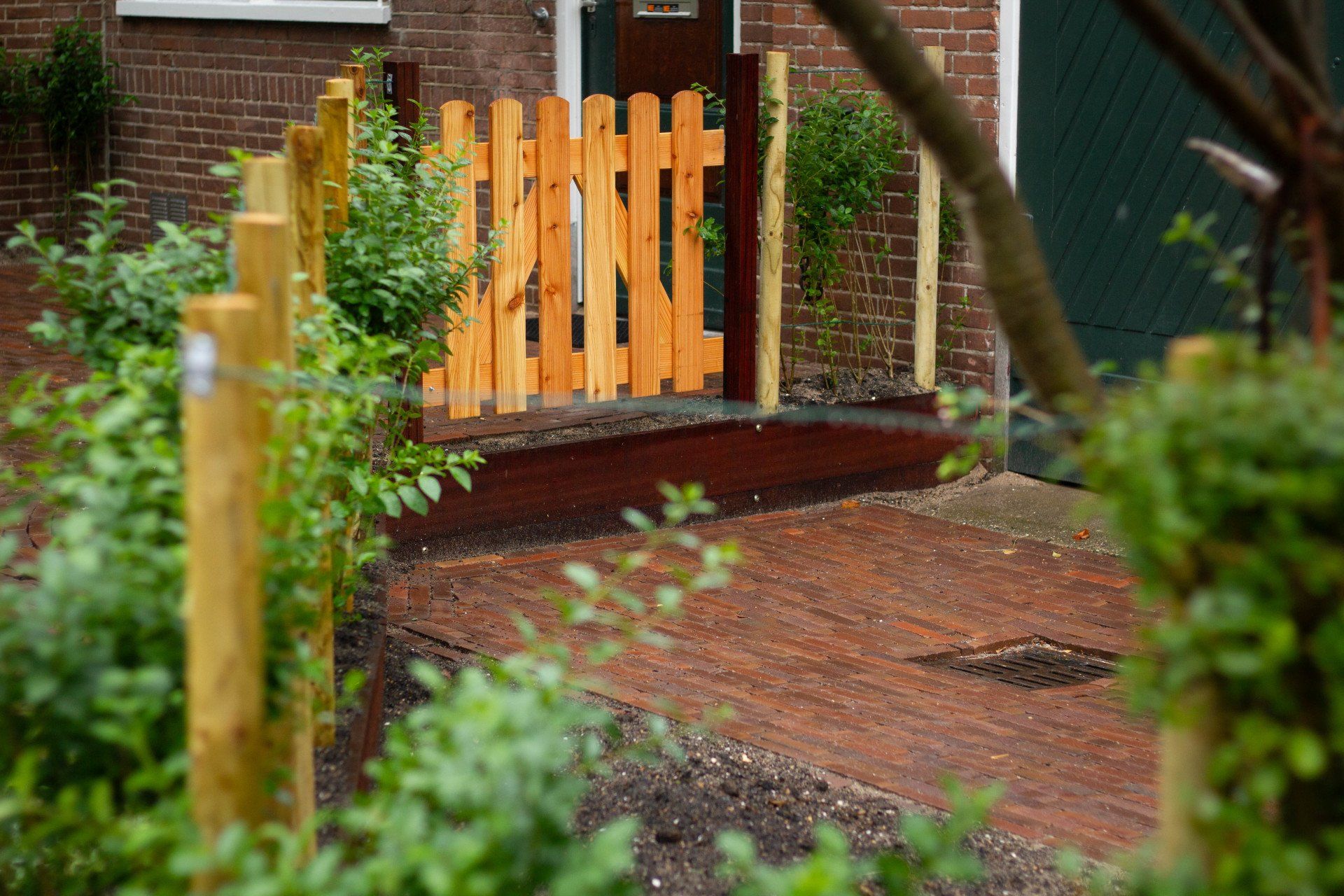 Houten poort voor een bakstenen gebouw. ​​Houten hekpalen markeren de omtrek van een bakstenen pad, met groen op de voorgrond.