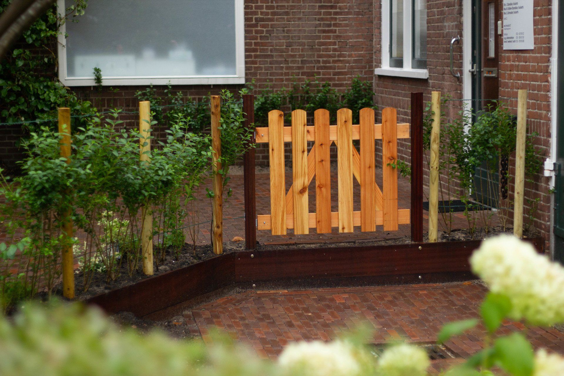 Houten poort in een kleine tuin, met een bakstenen huis en planten op de achtergrond.