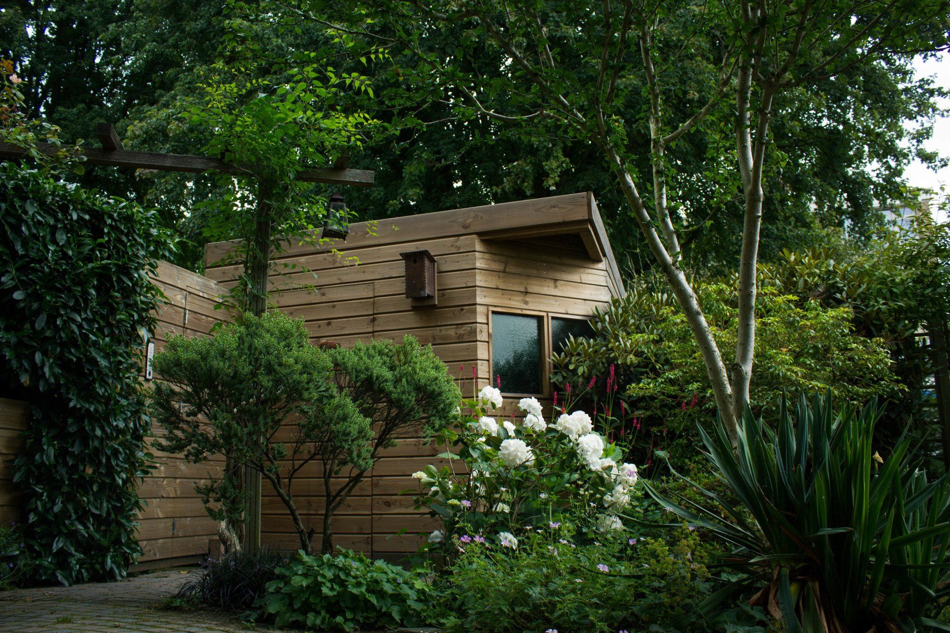 Een klein gebouw met bruine muren, verscholen in een weelderige, groene tuin vol bloemen en bomen.