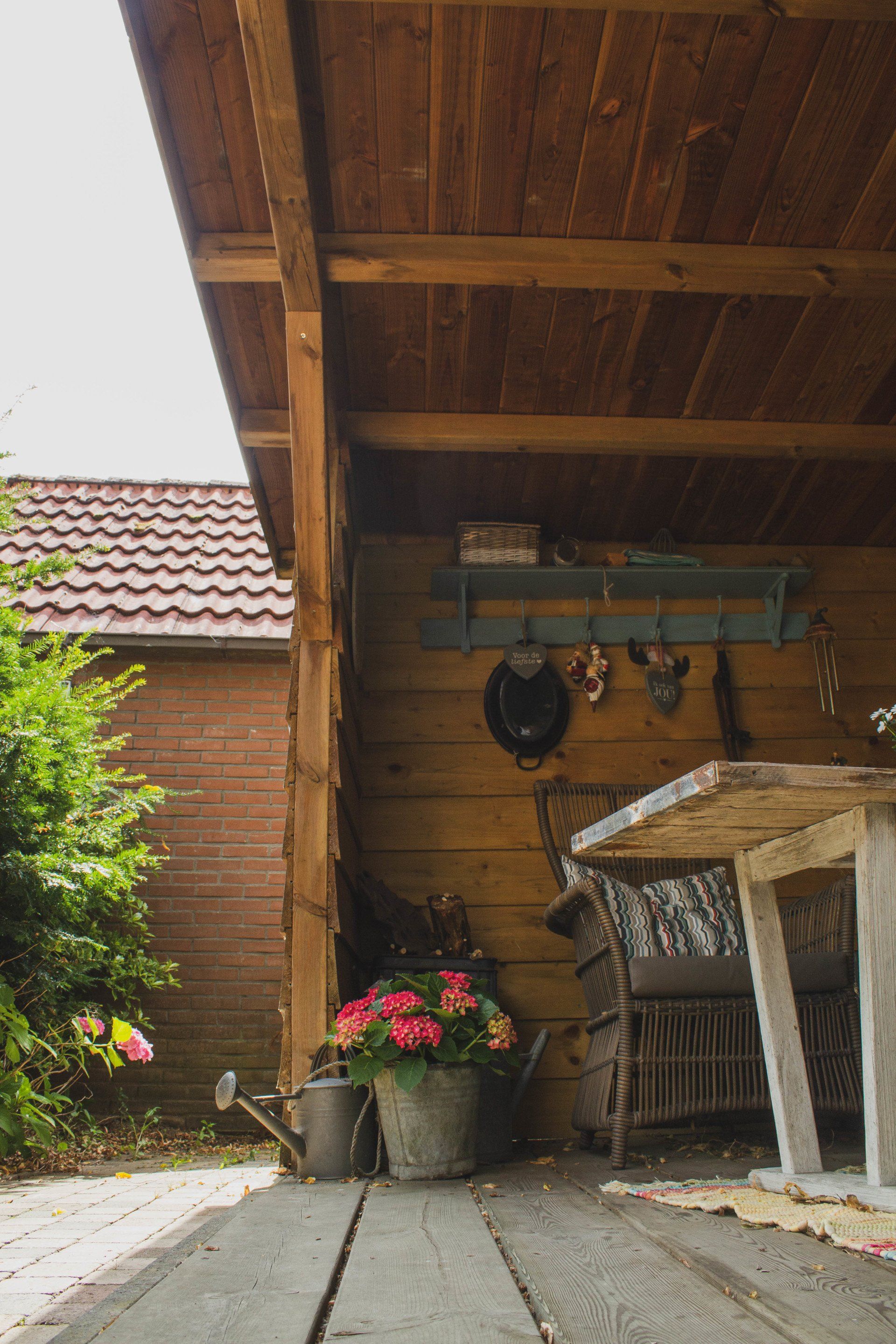 Houten schuur met tafel, bloemen en gieter. Op de achtergrond een rood pannendak.