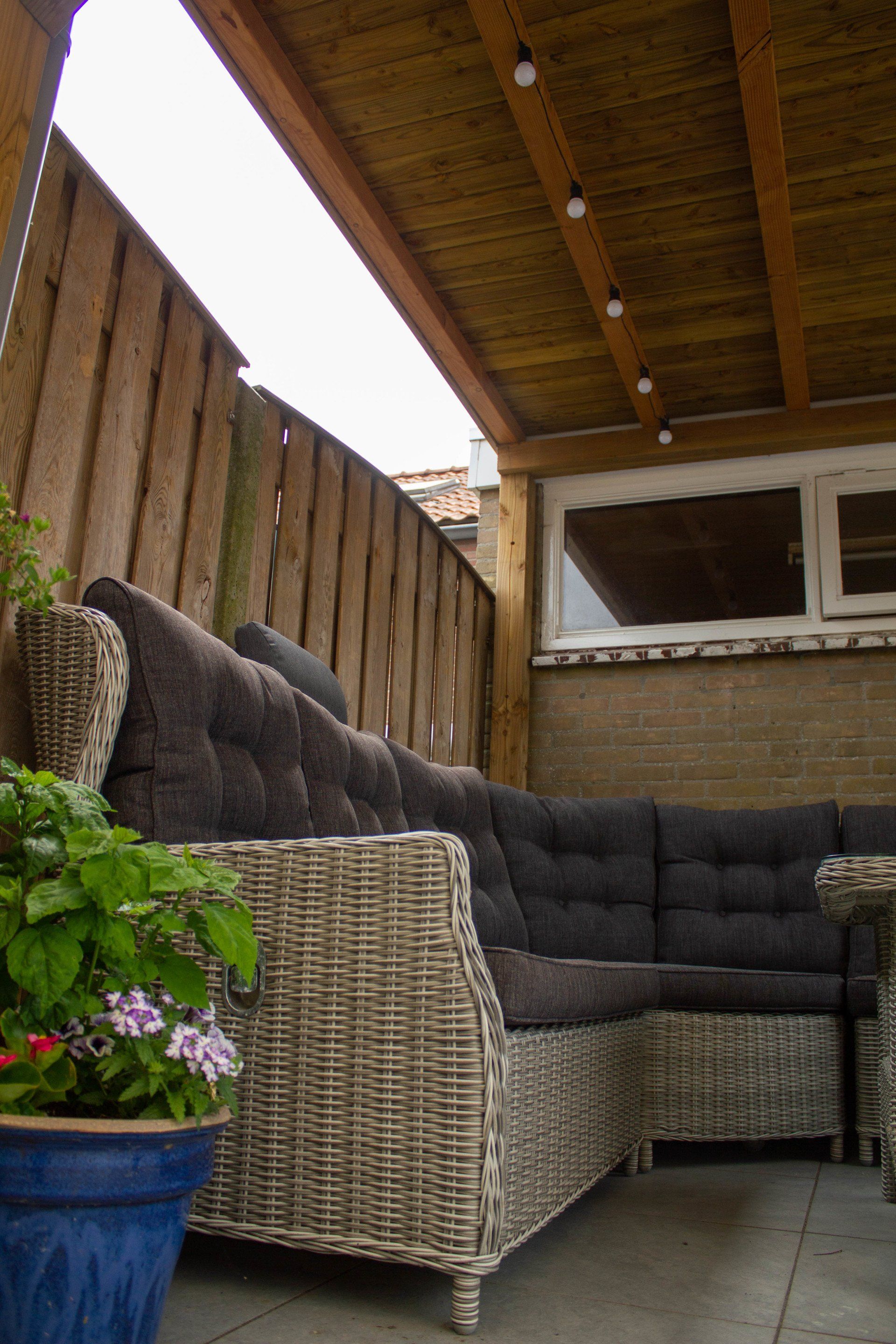 Een modulaire loungeset van riet met donkere kussens voor buiten, onder een houten afdak, naast een bloempot en een schutting.